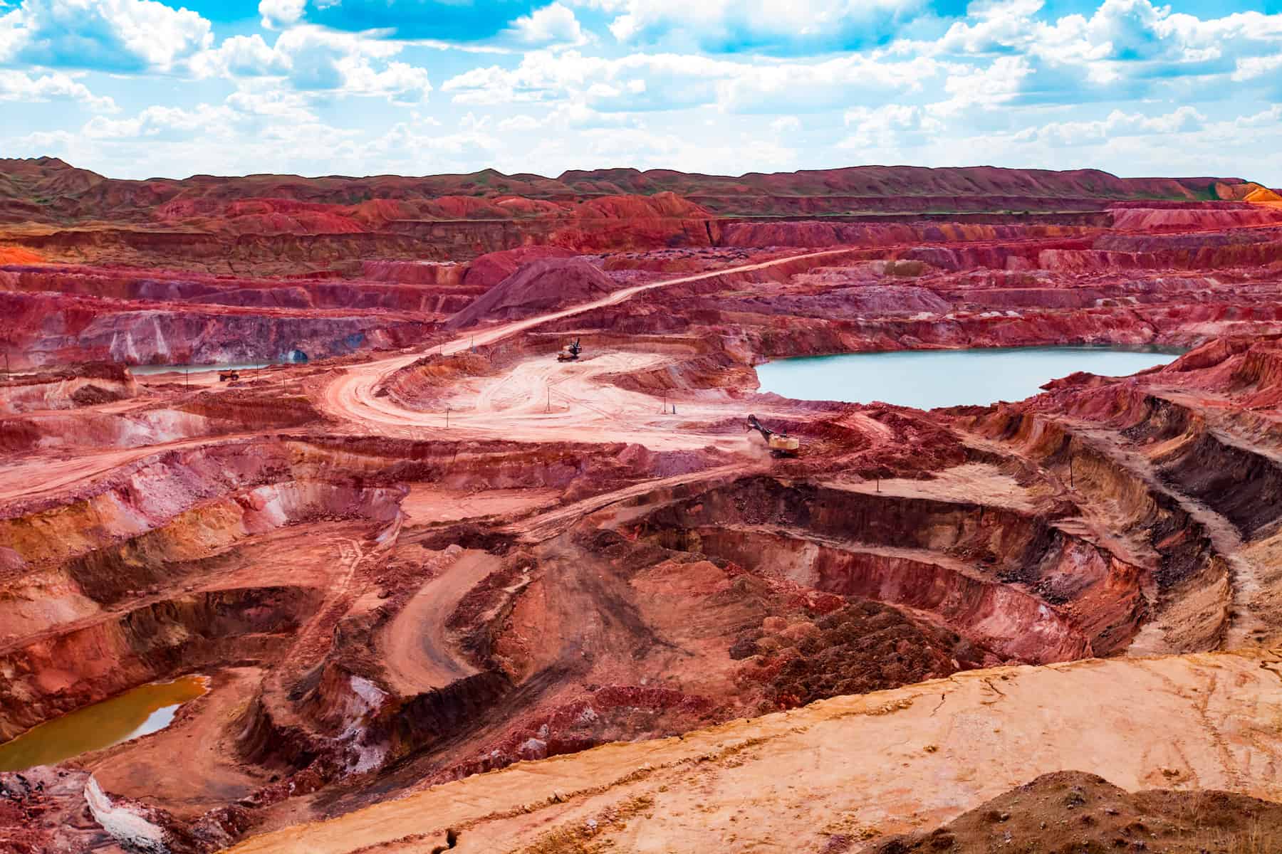 Manufacturer's aluminum ore quarry with excavators, dump trucks, and blue lake under a cloudy sky.