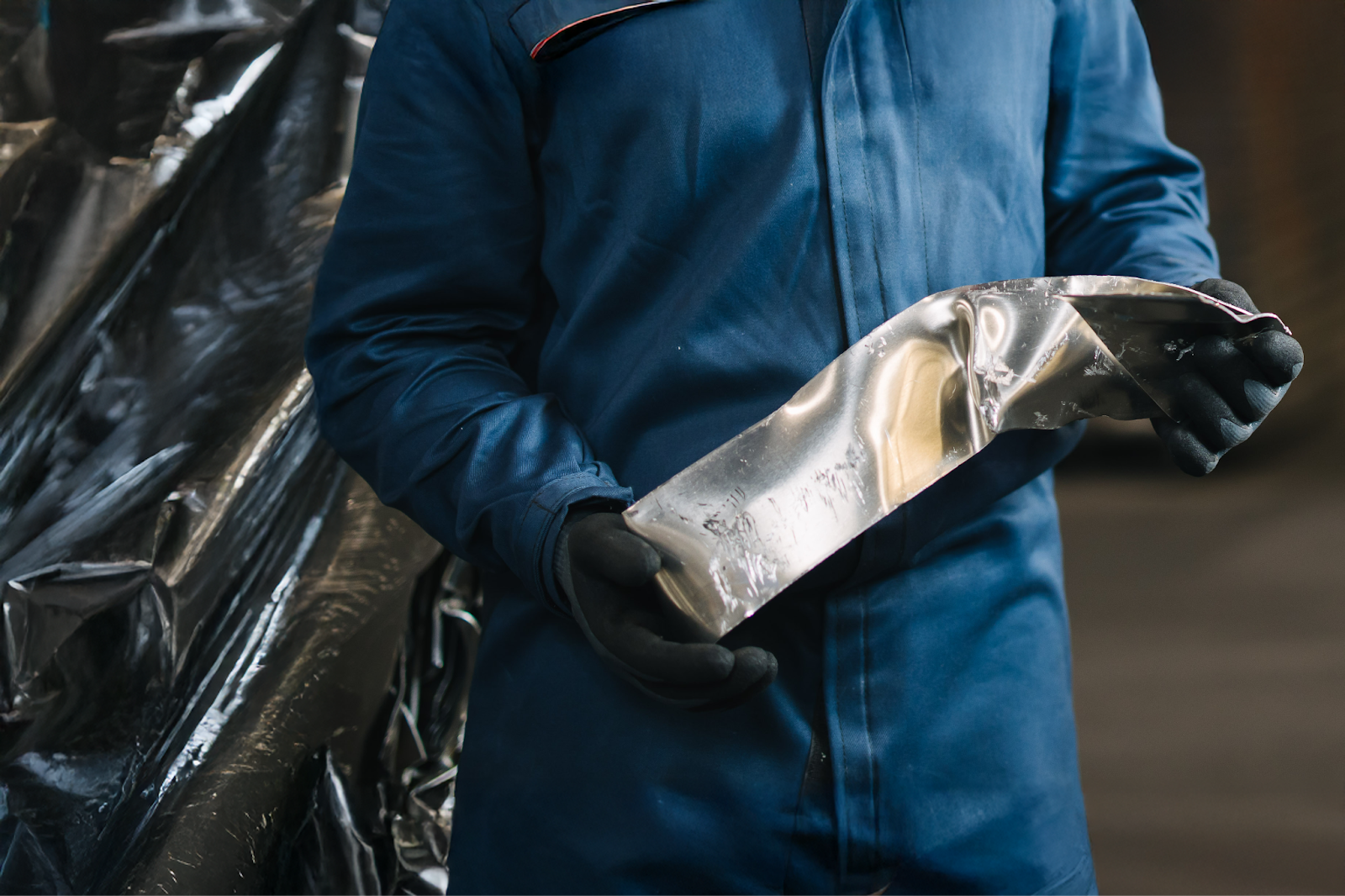 Worker in blue coveralls holding a bent aluminum strip sample for inspection, wearing gloves.