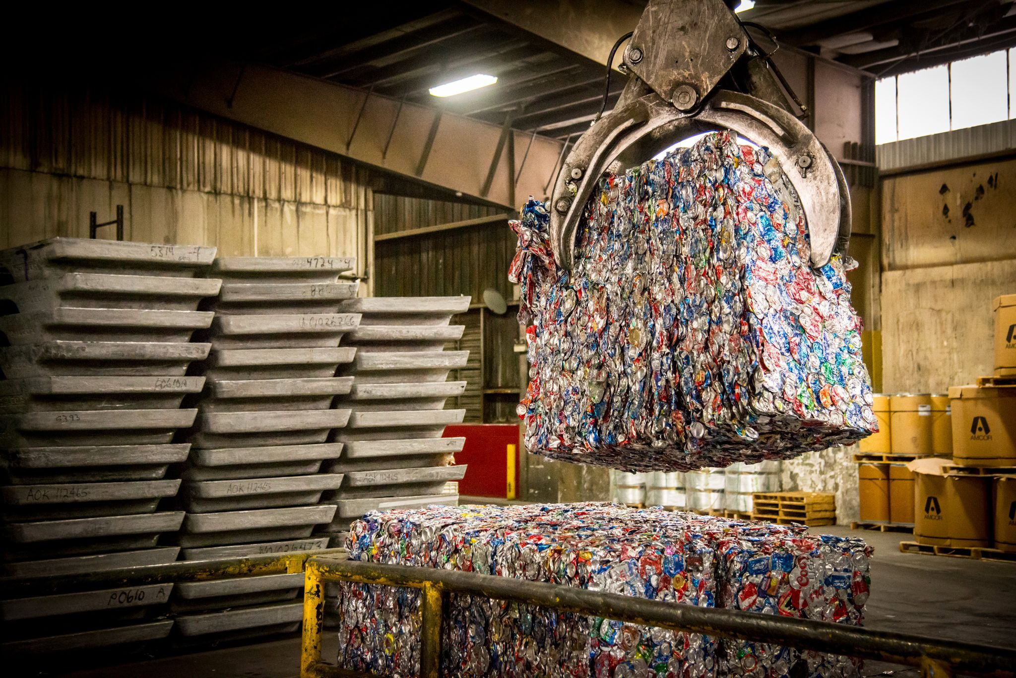 Industrial claw lifting a bale of crushed aluminum cans beside stacked ingots at a recycling plant