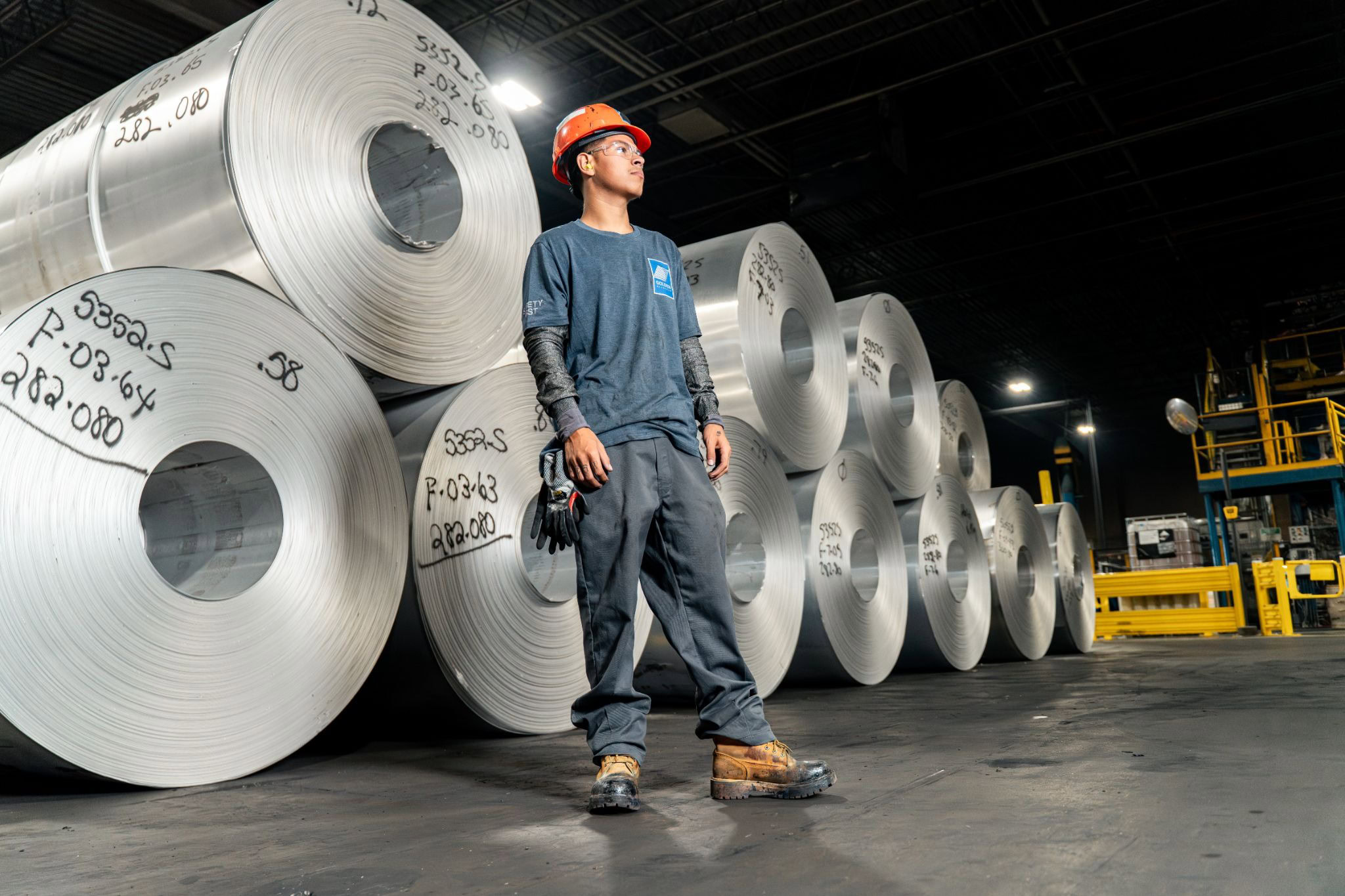 Worker in a hard hat standing among large rolls of flat-rolled aluminum in a mill