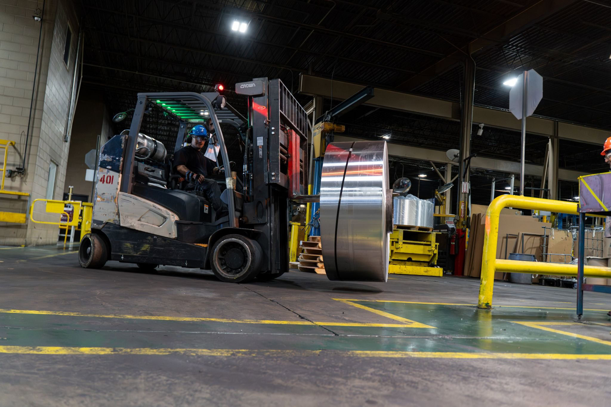 Forklift operator carries a large aluminum coil across the mill floor, past yellow safety railings and floor markings.