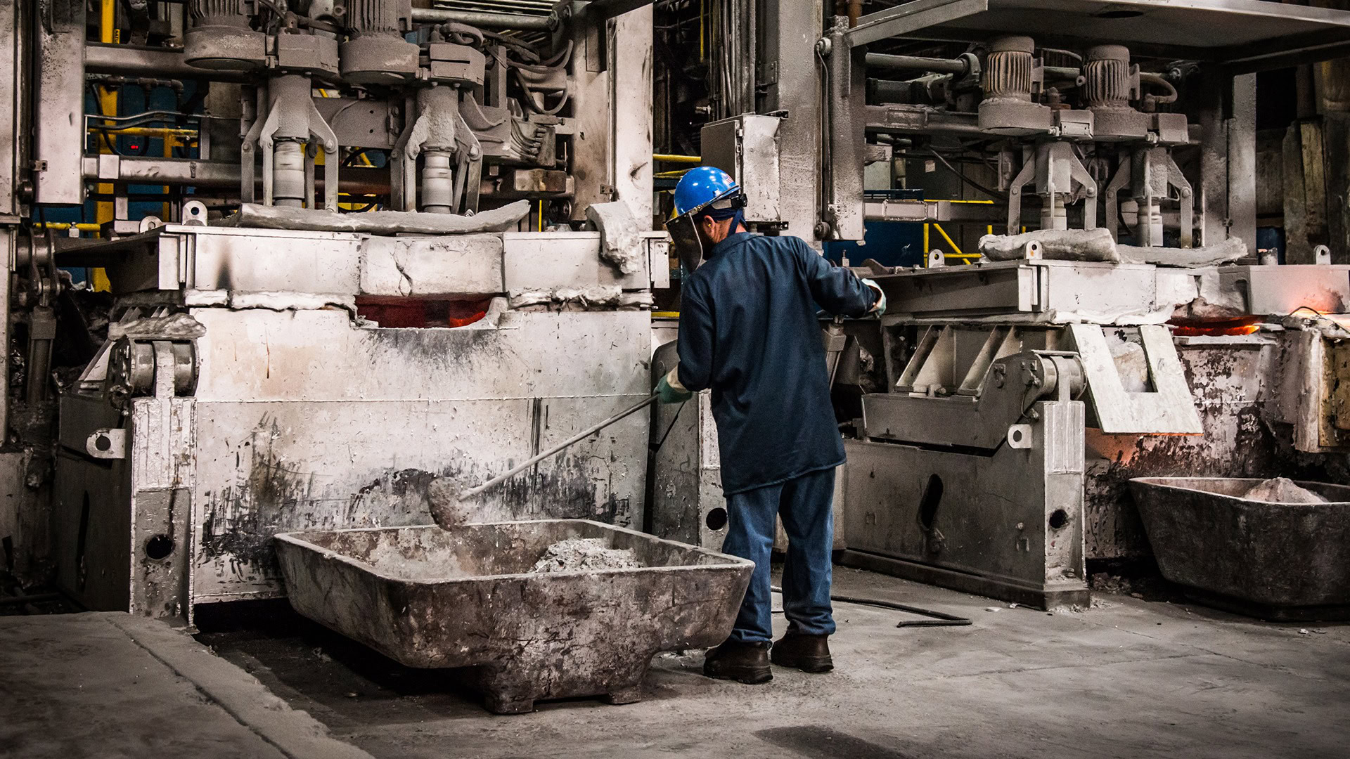 Worker in a hard hat and face shield tends a melting furnace, with a tray of scrap in the foreground.