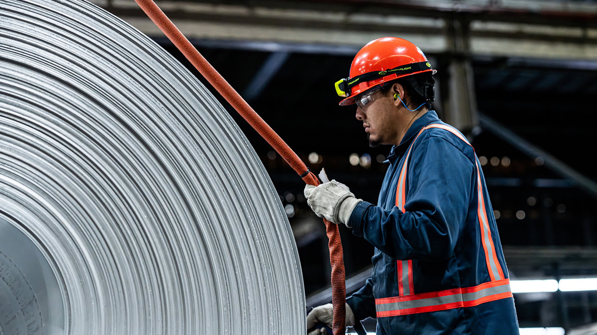 Worker in an orange hard hat and high-visibility vest handles a strap beside a large coil of aluminum.