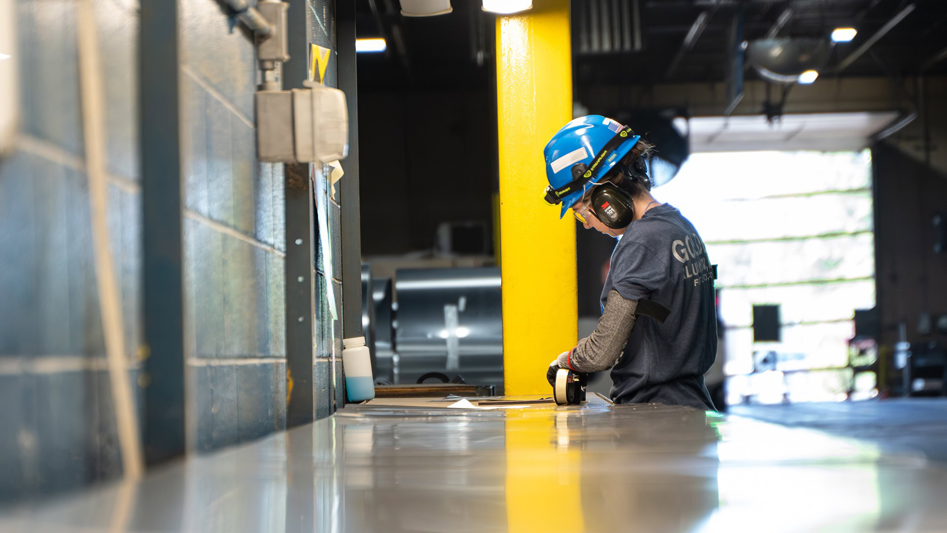 Worker in a hard hat and ear protection leans over a workstation during a final quality check before shipping.