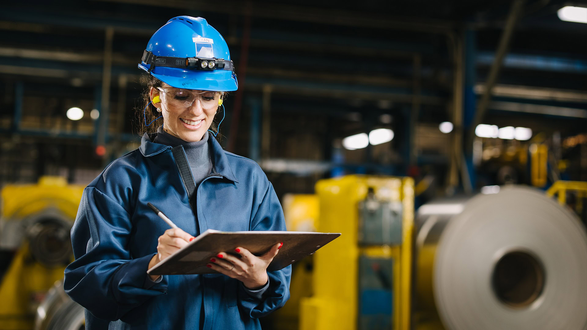 Smiling worker at Golden Aluminum takes notes on a clipboard in front of aluminum coils with hard hat and clipboard.